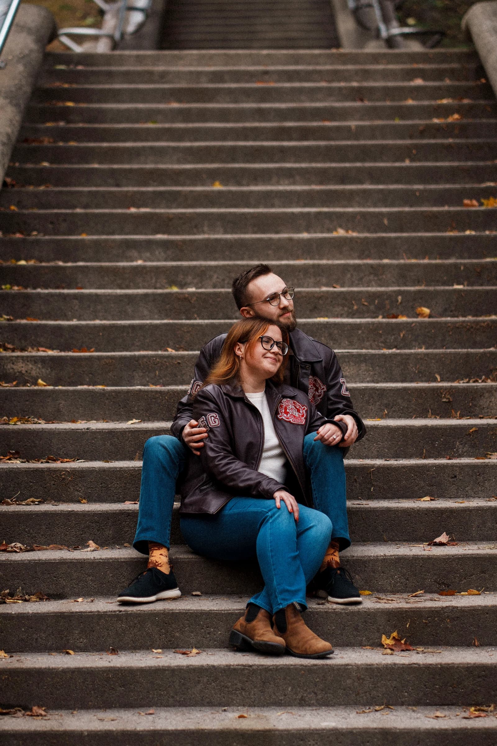 Rachel and Reid in leather jackets, sitting together on a wide stone staircase scattered with autumn leaves