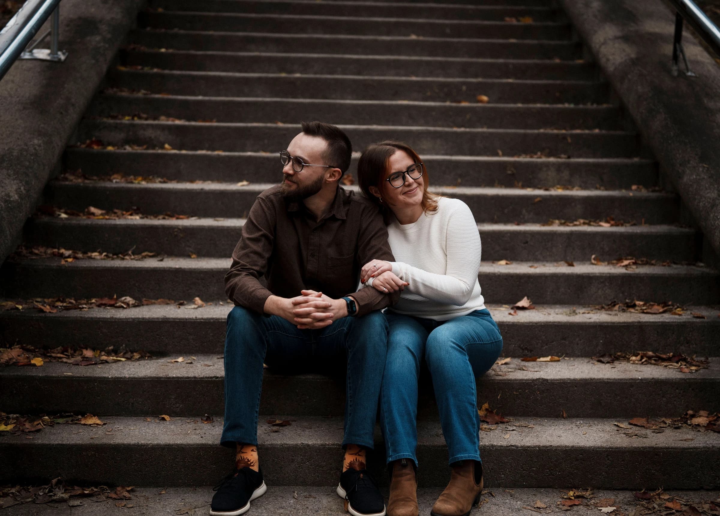 Rachel and Reid in leather jackets, sitting together on a wide stone staircase scattered with autumn leaves