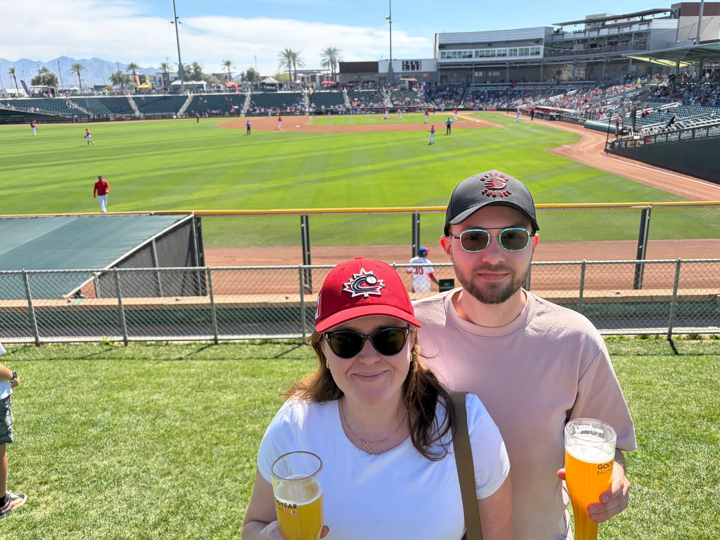 Rachel and Reid at the outfield fence at Goodyear Ballpark, beers in hand, with the game underway behind them